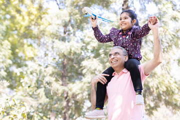 Senior man with granddaughter sitting on shoulders having fun flying toy airplane at park