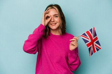 Young caucasian woman holding a English flag isolated on blue background excited keeping ok gesture on eye.