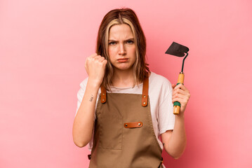 Young gardener woman holding gardening shovel isolated on pink background showing fist to camera, aggressive facial expression.