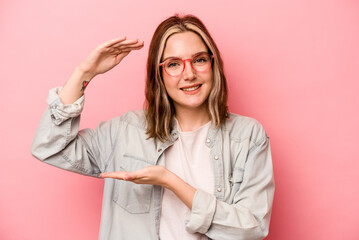 Young caucasian woman isolated on pink background holding something little with forefingers, smiling and confident.