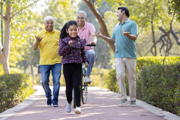 Obraz premium Senior man riding bicycle with granddaughter running while other old man and son encouraging at park
