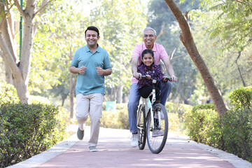 Cheerful senior man riding bicycle with granddaughter while son running at park