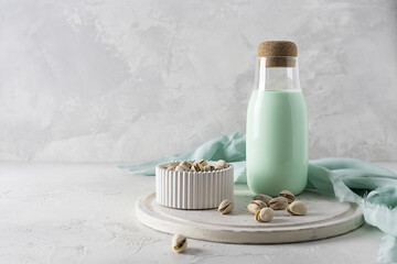 A glass bottle with pistachio milk on a light board. Pistachios in a white vase in the background. Light green napkin on the table. Light background, space for text