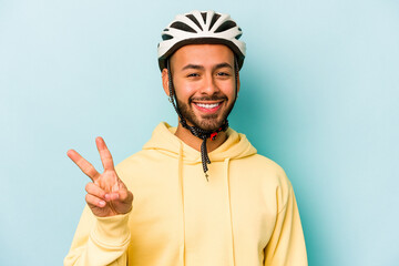 Young hispanic man wearing helmet isolated on blue background joyful and carefree showing a peace symbol with fingers.