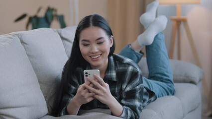 Asian female relaxing girl woman lying on couch at home enjoy chat typing app mobile phone relax watch viral live video play game follow like post in social media share and send smile emoji scroll web