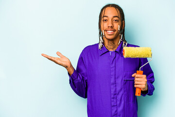 Young African American painter man isolated on blue background showing a copy space on a palm and holding another hand on waist.