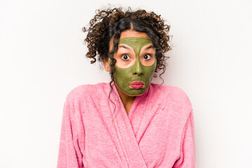 Young hispanic woman wearing a facial mask isolated on white background shrugs shoulders and open eyes confused.