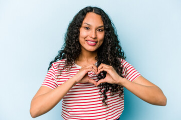 Young hispanic woman isolated on blue background smiling and showing a heart shape with hands.