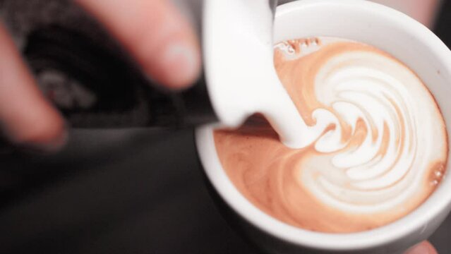 barista hands making latte art cappuccino pouring frothed milk from pitcher into white ceramic mug with espresso coffee in ultra slow motion closeup
