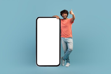 Positive indian guy posing by huge smartphone with blank screen