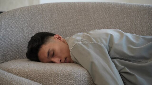 Tired Asian man falls down on couch falling asleep. Exhausted young male person wants to relax in living room after hard working day close view