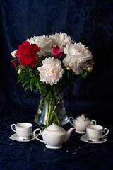 White tea-set and a glass vase with colourful peonies on the dark blue background.