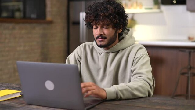 Happy and optimistic Indian man in casual wear using laptop in modern flat. Successful inspired Arabic male entrepreneur looks away and smiling thoughtfully, planning next step