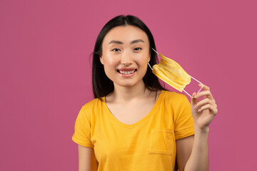 End of coronavirus pandemic. Happy asian lady taking off medical mask, feeling free over violet studio background