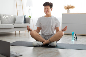 Asian man meditating in lotus position on mat with pc