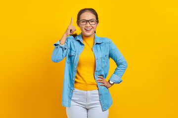 Portrait of smiling young Asian woman student in denim clothes pointing finger in copy space, showing advertising products isolated on yellow background. Education in college university concept