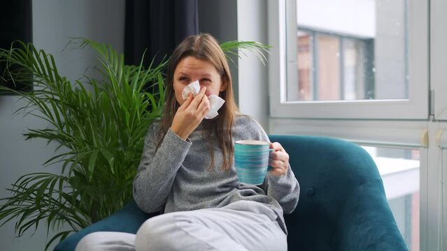Unhealthy woman sits in a chair, drinks hot tea or a cold medicine and sneezes or blows her nose into a napkin because she has a cold, flu, coronavirus. It is snowing outside