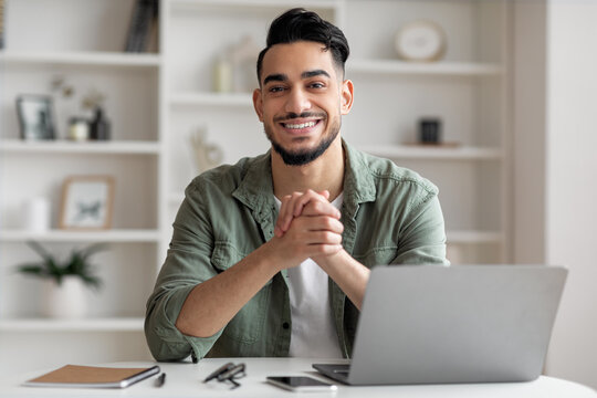 Glad handsome young islamic man with beard at workplace with laptop in home office interior