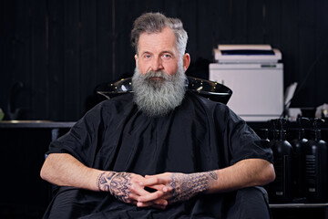 Portrait of cool old man with tattoos and long beard sitting on armchair in hair salon.