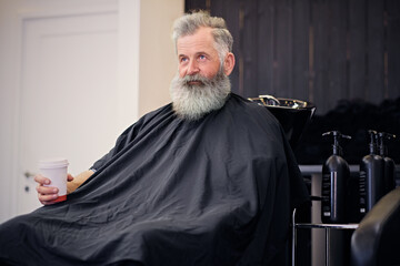 Shot of long bearded old hipster with black cape on armchair in barbershop with stylish interior.