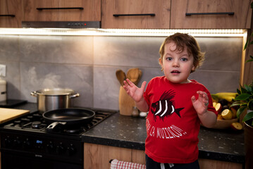 Zdziwiony chłopiec w kuchni, w tle gotuje się obiad, na kuchence stoi parujący garnek. Mały kucharz w kuchni.