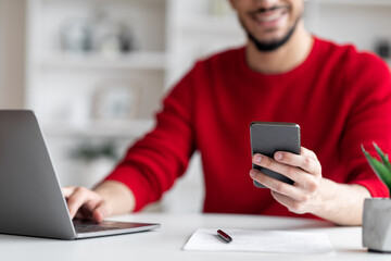 Happy young arab male with beard in red clothes typing on smartphone and laptop at workplace in...
