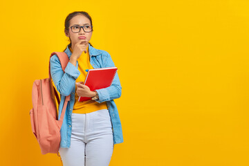 Portrait of pensive young Asian woman student in casual clothes with backpack holding book and thinking about question isolated on yellow background. Education in college university concept