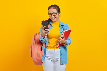Portrait of cheerful young Asian woman student in denim clothes, glasses with backpack holding mobile phone and books isolated on yellow background. Education in high school university college concept