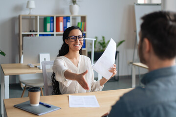 Happy HR manager holding CV, offering handshake to job applicant during employment interview at modern office