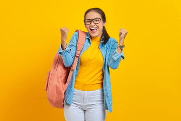 Cheerful young Asian student in denim outfit with backpack standing doing winning gesture...