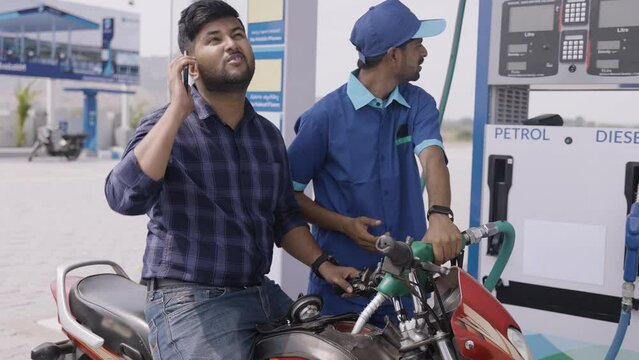 Customer busy using mobile phone while worker refuling petrol to bike at gas station - concept of safety, danger of using mobile pump and social media addiction.
