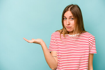 Young caucasian woman isolated on blue background showing a copy space on a palm and holding another hand on waist.