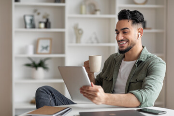 Smiling attractive young muslim male with beard in wireless headphones drink coffee, make online call