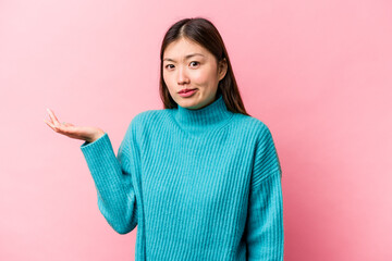 Young Chinese woman isolated on pink background doubting and shrugging shoulders in questioning...