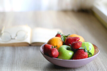 Bowl of apples, oranges and strawberries, open book and reading glasses on the table. Selective focus.