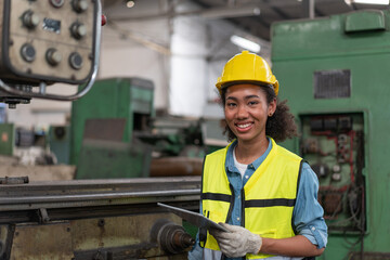 Portrait of female engineer in safety vest with yellow helmet smiling and holding tablet in old machine background at factory Industrial