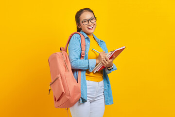 Smiling young Asian woman student in denim clothes with backpack, writing notes in notebook...