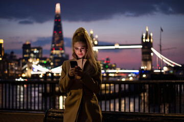 A pretty city woman looks at her smartphone during night time in front of the illuminated skyline...