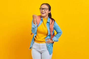 Cheerful young Asian woman student in denim outfit with backpack standing doing winning gesture celebrating isolated on yellow background. Education in university concept