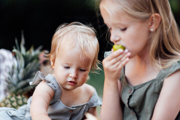 Candid lifestyle portrait of two caucasian siblings eating healthy food on picnic blanket at summer. Cute baby one year old and her sister seven years old having fun together during vacations on