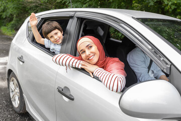 Happy arabic wife in hijab and child waving hands in open window at car, husband at steering wheel