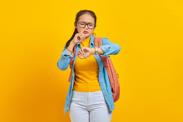 Confused young Asian woman student in denim outfit with backpack checking time on watch isolated on yellow background. Education in university college concept