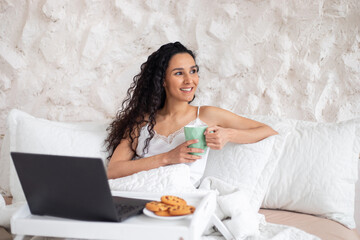 Obraz na płótnie Canvas Happy millennial caucasian brunette female with long hair in pajamas have breakfast and drinks coffee on bed