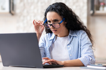 Eyesight problems. Latin lady using glasses while working on laptop computer at home, sitting in...