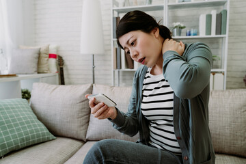 chinese millennial woman tries to relive shoulder tension while looking at her phone screen. poor sitting posture is causing neck pain to the pretty asian lady. healthcare and medical concept - obrazy, fototapety, plakaty