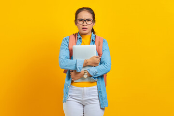 Portrait of surprised young Asian woman student in casual clothes with backpack holding laptop and looking at camera isolated on yellow background. Education in university college concept
