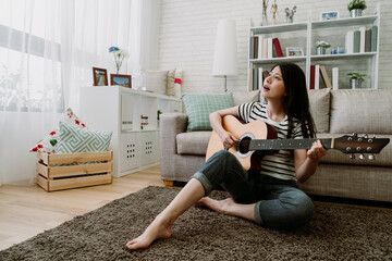 asian female composer sitting on living room carpet is making music with guitar. pretty taiwanese girl playing and sing country songs in her free time. indoor recreation concept.