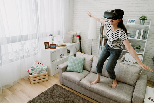 asian female player with outstretched hands hovering in the sky in virtual reality dreams. japanese woman standing on couch is experiencing fun of vr tech. future technology and indoor recreation