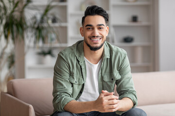 Smiling attractive millennial middle eastern guy blogger with beard looks at camera, sits on sofa...