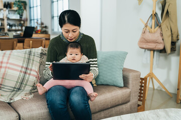asian mother and adorable baby are sitting in living room and watching pictures on tablet. chinese female parent and her kid are viewing fun videos online with a pad on sofa. - Powered by Adobe